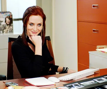 A person sitting at a desk in an office, smiling, with a red filing cabinet and magazines visible in the background.