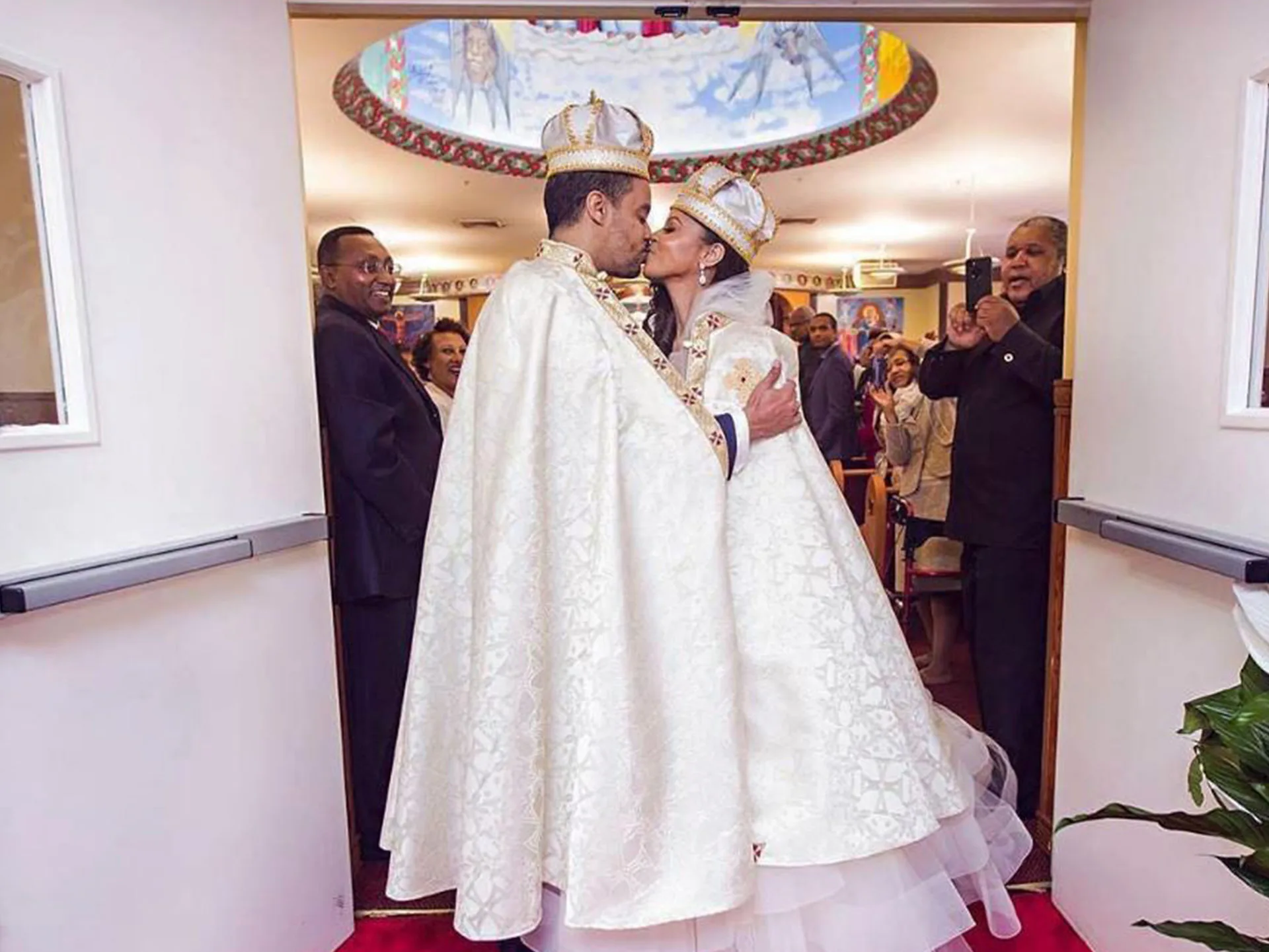 A couple in ornate robes and crowns kiss at an Ethiopian-style wedding ceremony, surrounded by guests.