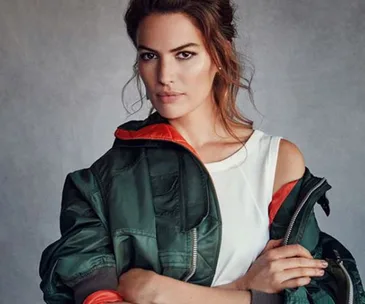 A woman in a green jacket and white top stands with a confident expression, her arms crossed.