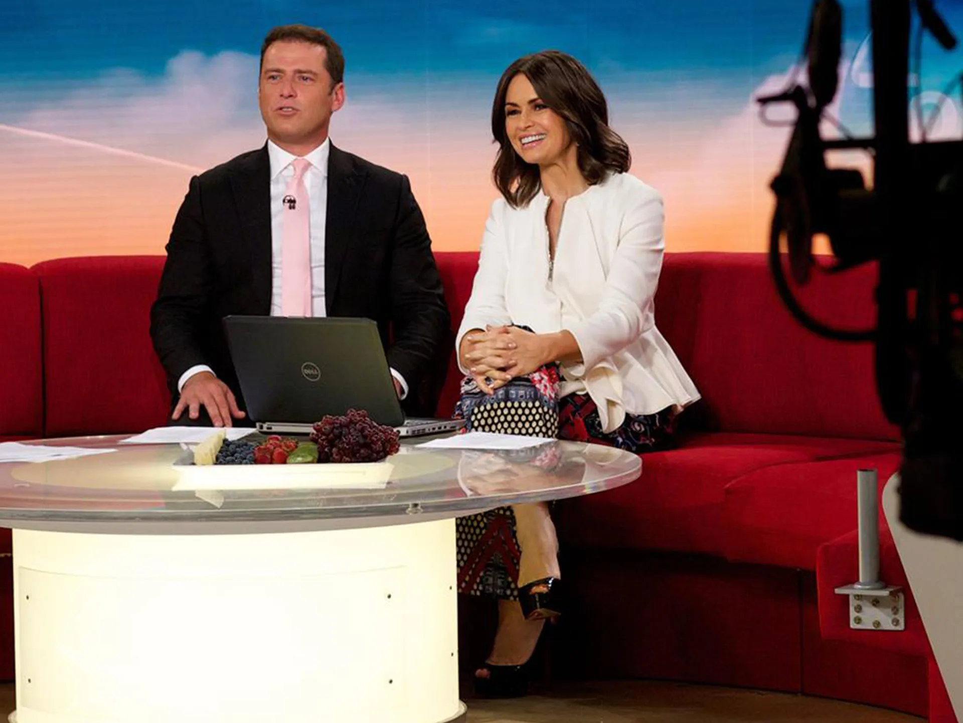 Two TV hosts sitting on a red couch with a laptop on the table in a studio setting.