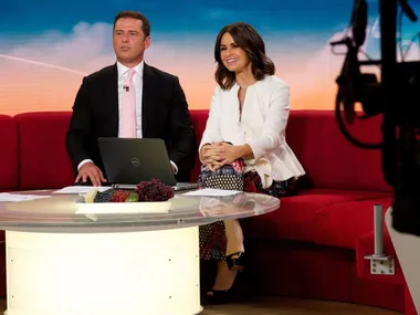 Two TV hosts sitting on a red couch with a laptop on the table in a studio setting.