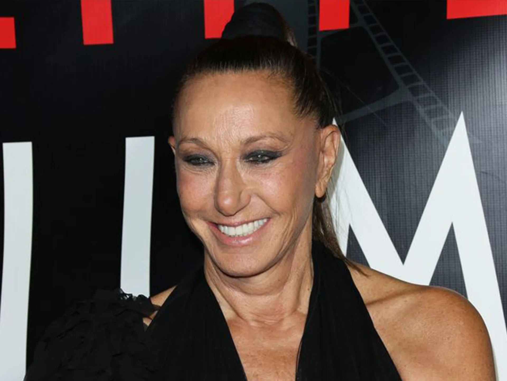 Donna Karan at an event, smiling, in a black halter top against a black and white background.