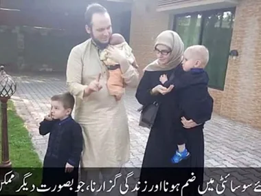 A family of four, including two young children, stand outside on a paved area. A subtitle in Urdu is overlaid at the bottom.