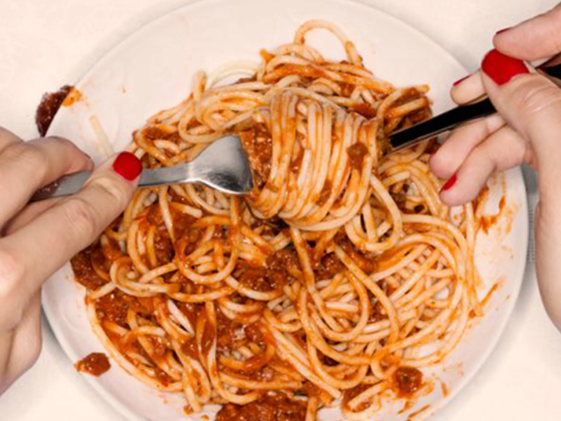 Hands twirling spaghetti with tomato sauce on a plate using a fork while stabilizing with a spoon.