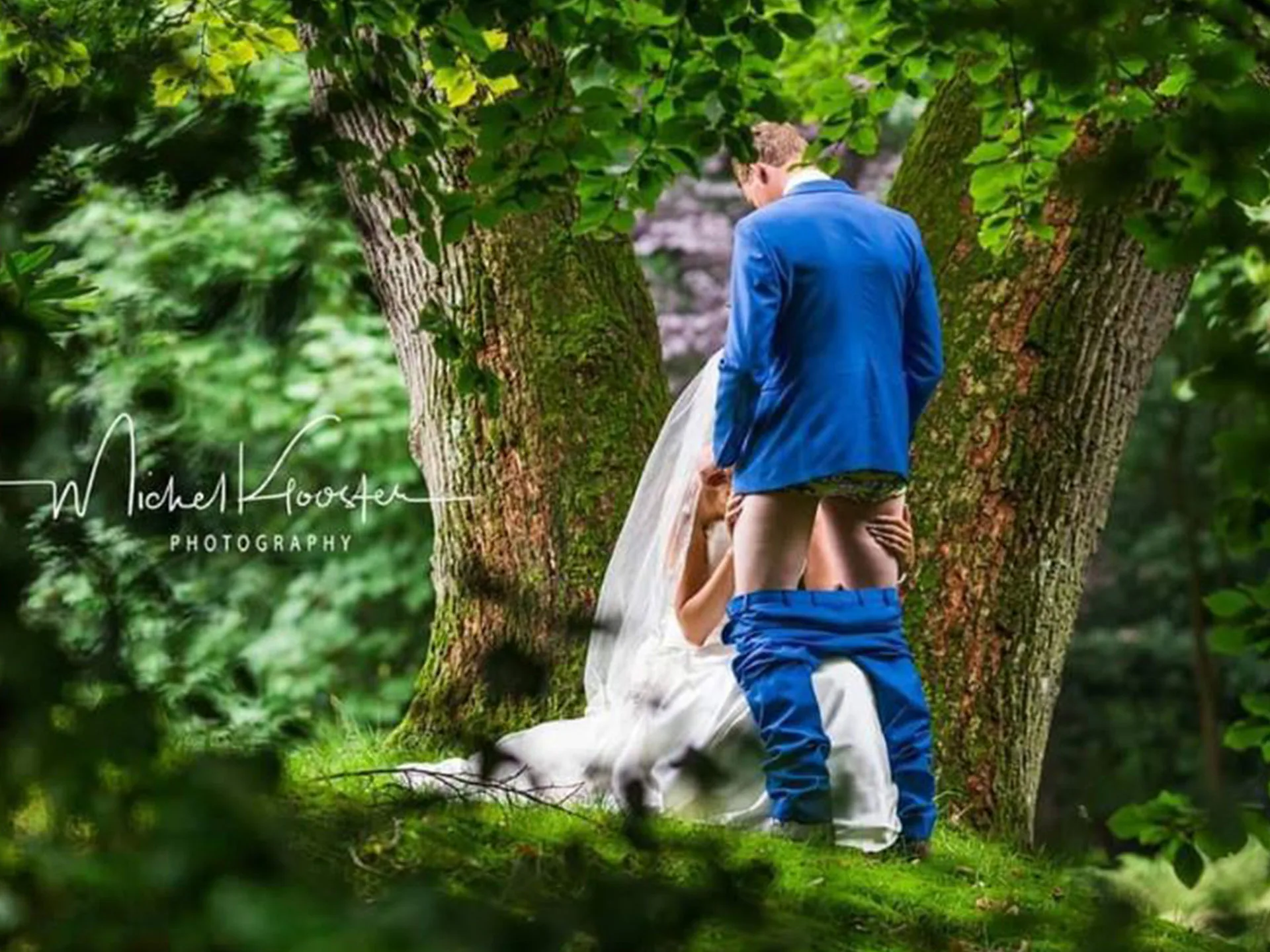 A bride kneels in front of a groom with his pants down, set in a forested area.