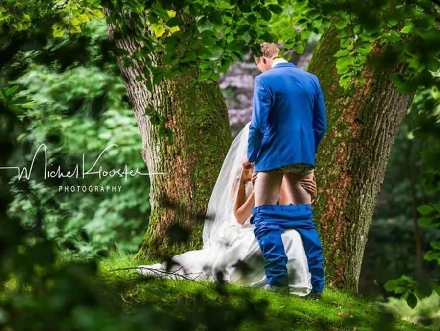 A bride kneels in front of a groom with his pants down, set in a forested area.