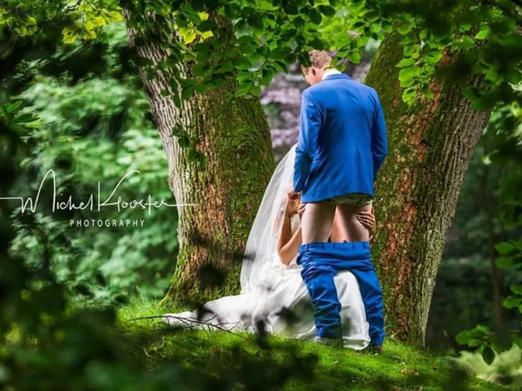 A bride kneels in front of a groom with his pants down, set in a forested area.