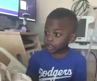 A young boy in a blue Dodgers shirt stands in a hospital room next to medical supplies and a computer.