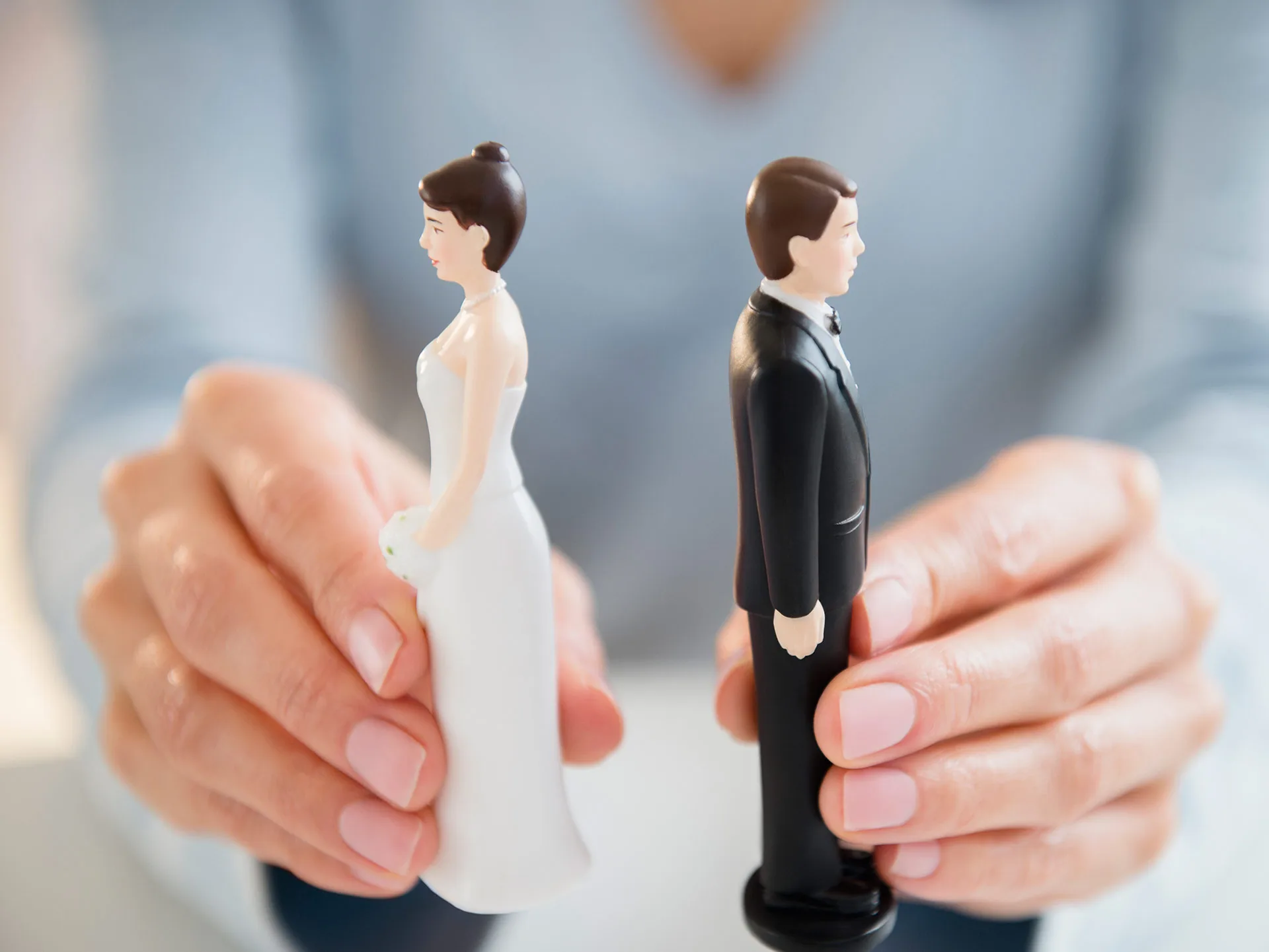 Hands holding wedding cake topper figurines of a bride and groom facing away from each other.