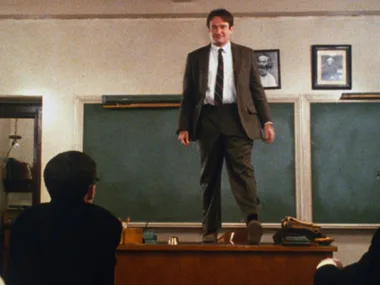 Teacher stands on a desk in a classroom, engaging students, with a chalkboard and framed photos in the background.