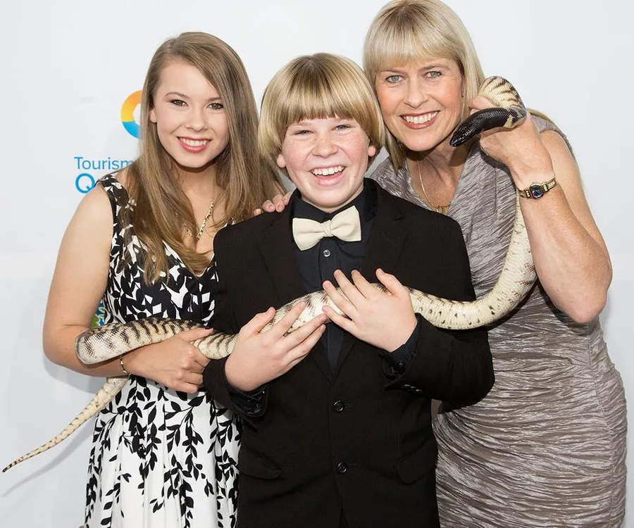 Three people smiling, holding a large snake, dressed formally at an event.