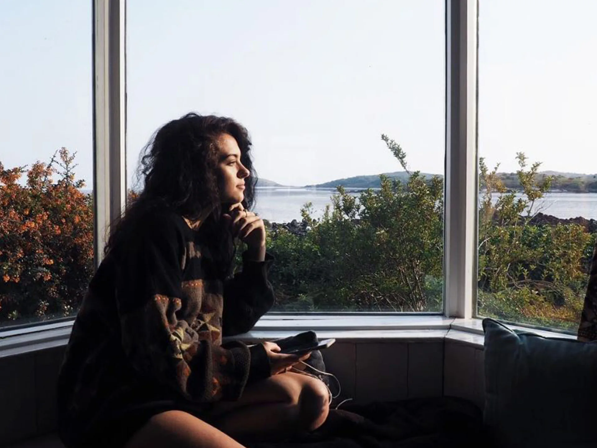 Woman sitting by a large window, looking at a scenic view of greenery and water, holding a phone.