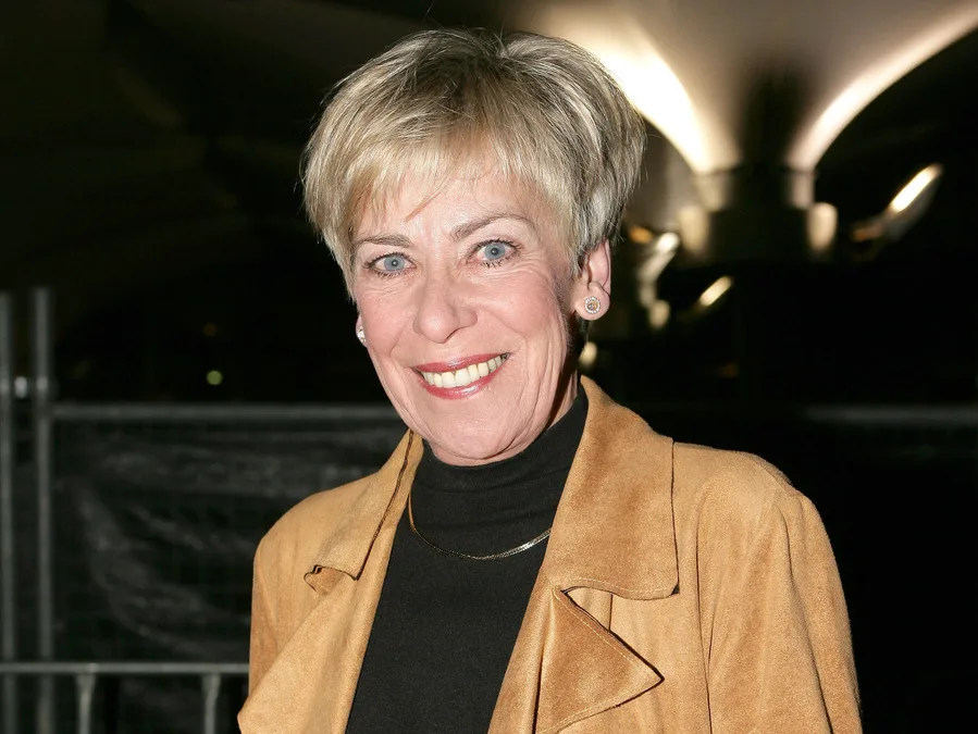 Smiling woman in a brown jacket and black top, with short blonde hair, in night setting.