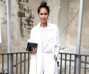 Woman in white outfit holding a black clutch, standing in front of a textured wall.