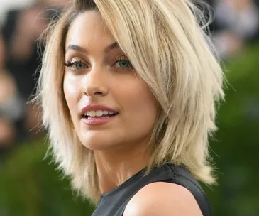 A woman with short blonde hair at an event, wearing a sleeveless black dress, looking over her shoulder.