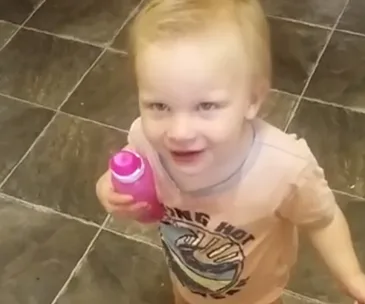 A toddler smiling and holding a pink bottle, standing on a tiled floor.