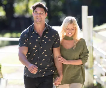 Man and woman walking outdoors, smiling, arm in arm, with lush greenery background.