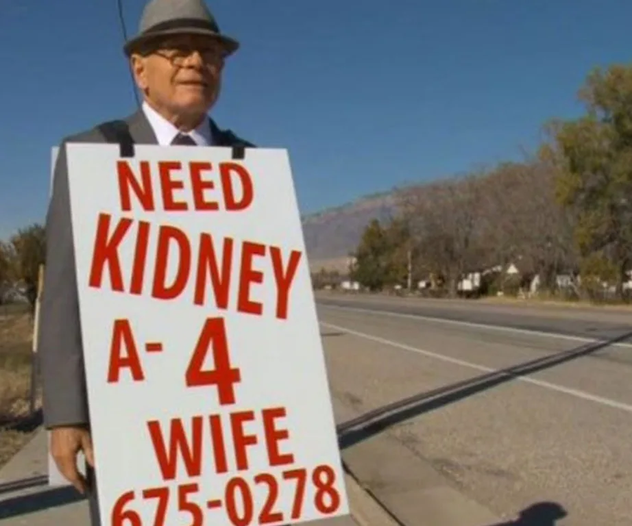 Man wearing suit and hat holds sign asking for a kidney donor for his wife with blood type and phone number.