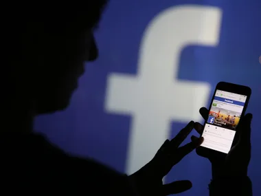 Silhouette of person using smartphone displaying Facebook app with a Facebook logo in the background.
