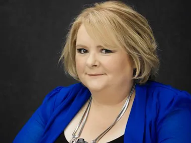 Alt text: "Person in a blue blazer with a short haircut, smiling against a dark background."