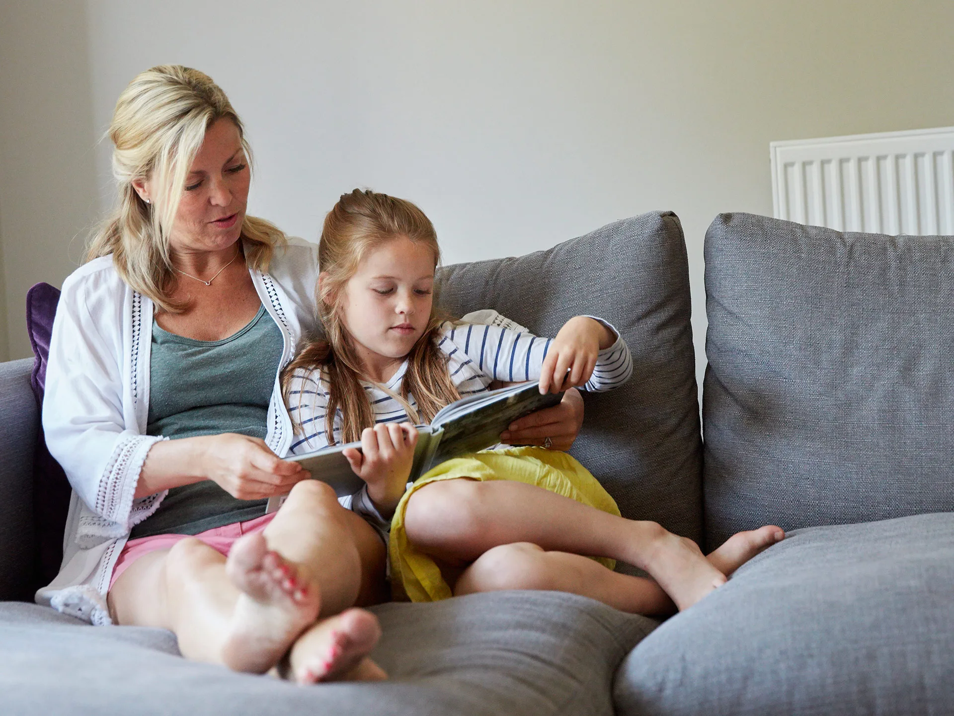 A woman and a young girl sit on a gray couch, reading a book together, sharing a quiet moment.