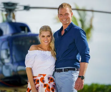 A smiling couple stands in front of a helicopter, dressed casually with tropical background.