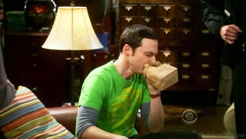 Sheldon Cooper from The Big Bang Theory hyperventilating into a paper bag in a dimly lit living room.