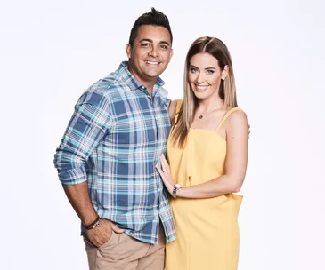 A couple stands together, smiling warmly; the man in a plaid shirt and the woman in a yellow dress.