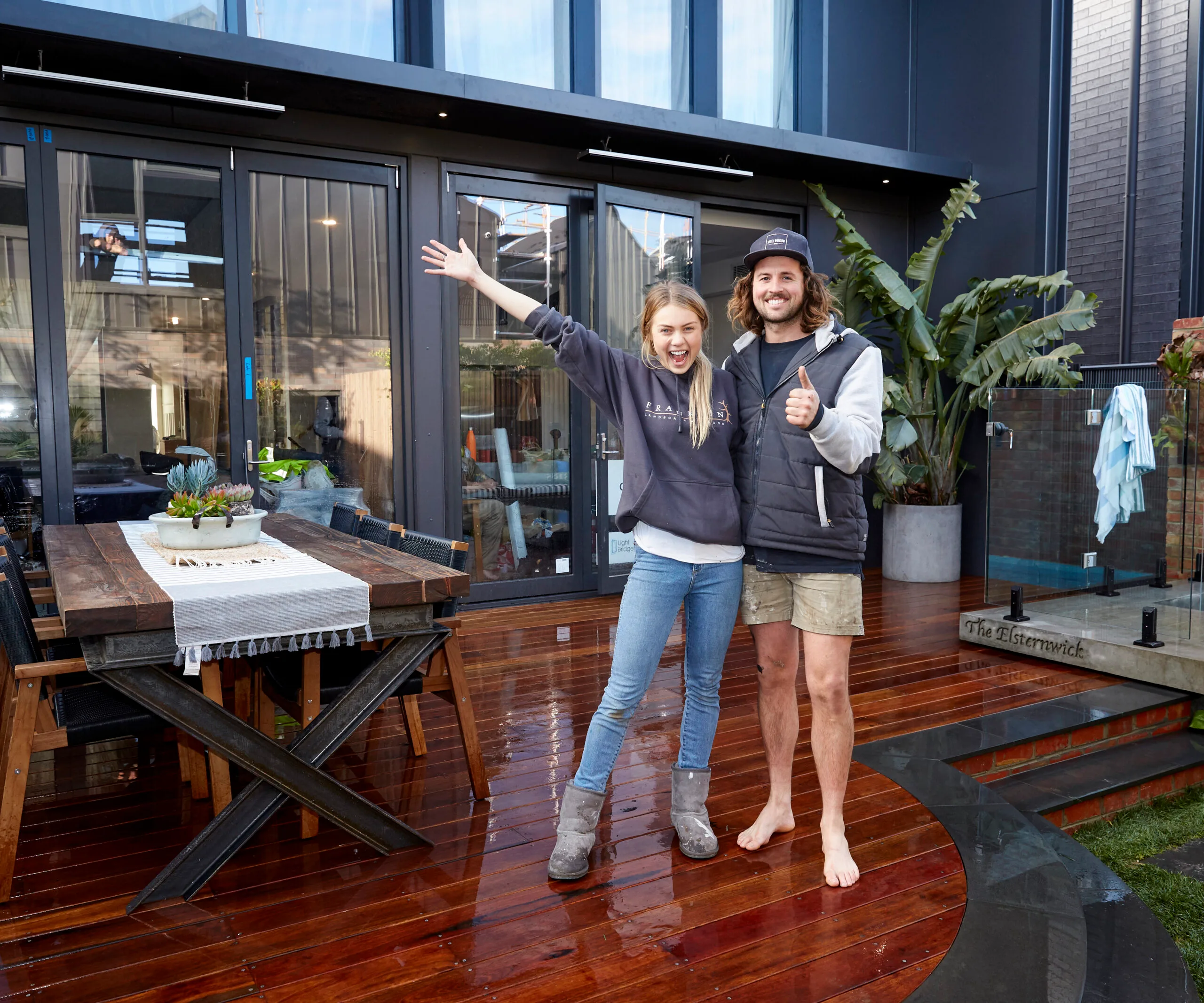 Couple posing excitedly on a wet wooden deck in front of a modern house, with outdoor dining table and plants.