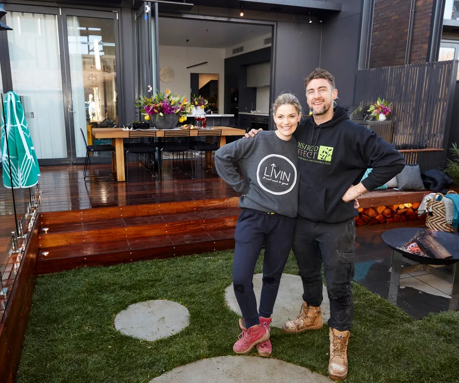 Couple smiling and standing in a modern backyard with a wooden deck, outdoor dining table, and fire pit in the background.