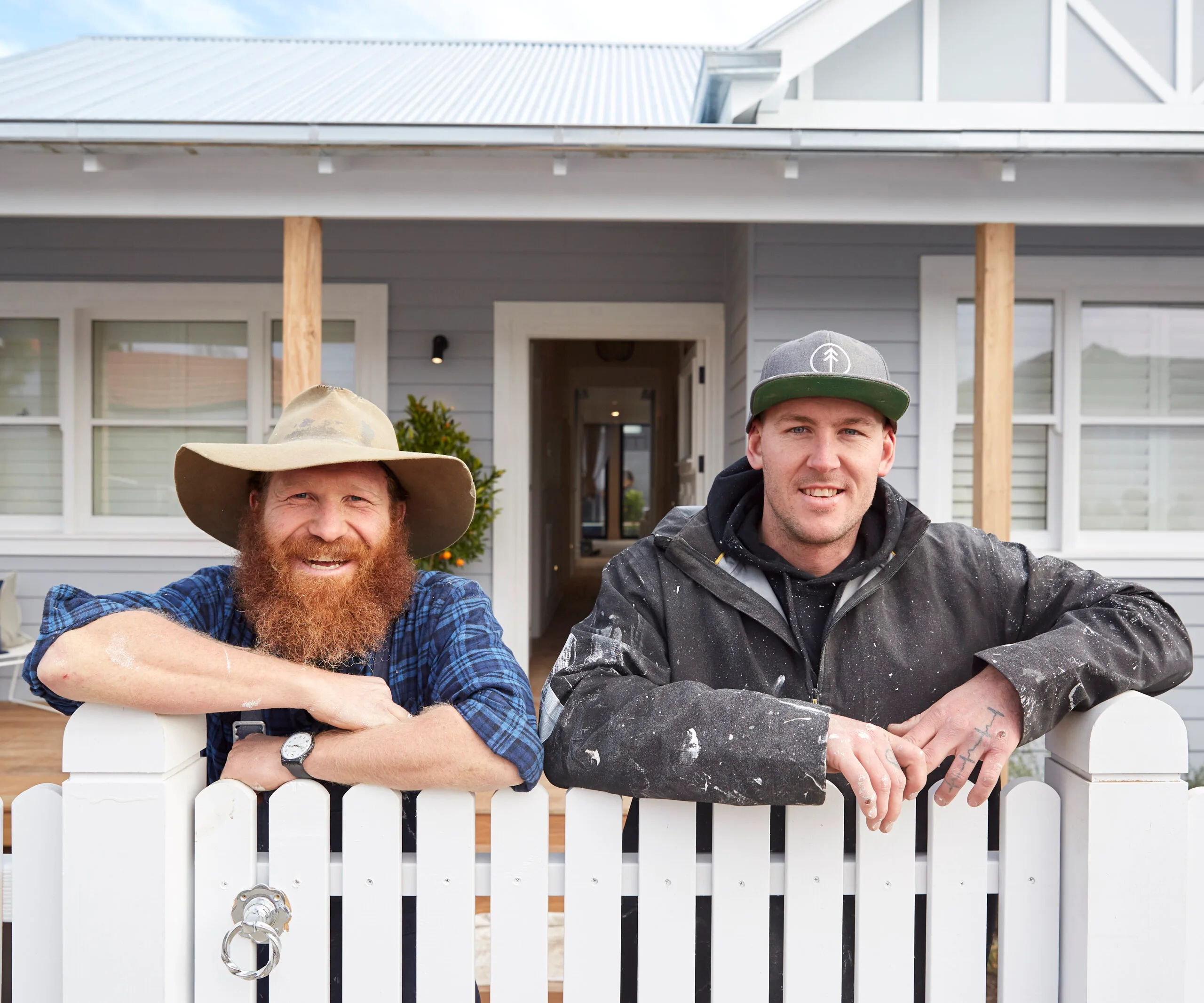 Two men smiling and leaning on a white picket fence in front of a grey house, one wearing a hat, the other a cap.