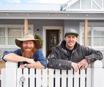 Two men smiling and leaning on a white picket fence in front of a grey house, one wearing a hat, the other a cap.