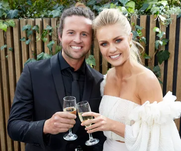 Smiling couple in formal attire holding champagne glasses, standing outdoors with leafy background.