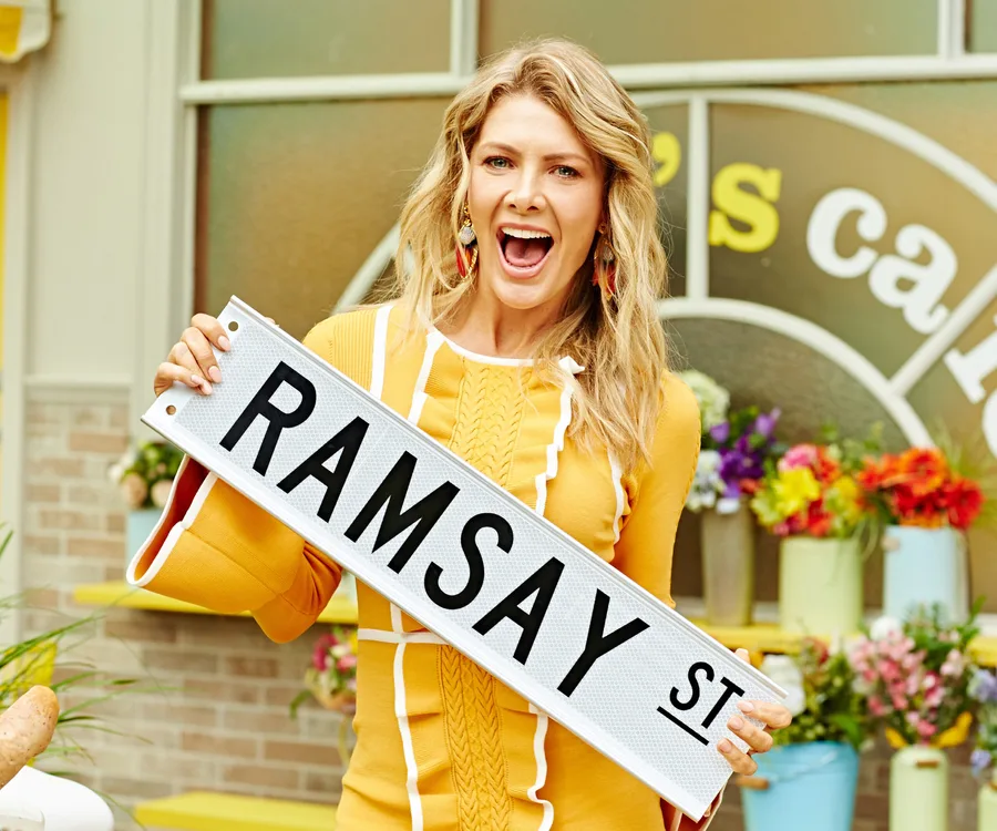 A woman in a yellow dress excitedly holds a "RAMSAY ST" sign, with colorful flowers in the background.