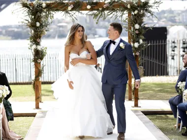 "Couple in wedding attire smiling, walking under floral arch outdoors."
