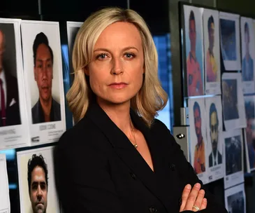 "Blonde woman in a dark blazer stands confidently in front of a board displaying various mugshots."