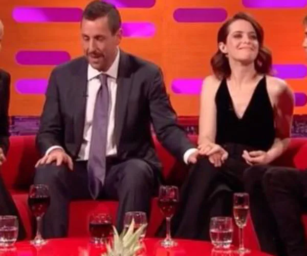 Graham Norton Show scene with two guests talking, one in a suit, the other in black, holding hands on a red sofa.
