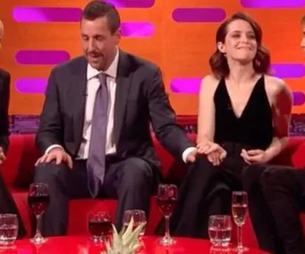 Graham Norton Show scene with two guests talking, one in a suit, the other in black, holding hands on a red sofa.