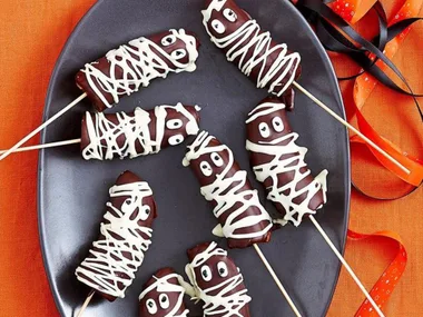 Mummy-themed chocolate cake pops with white icing drizzle and googly eyes on skewers, placed on an orange background.