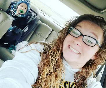 Young woman smiling in a car selfie with a child in a car seat in the back seat.