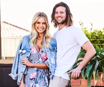 A smiling woman in a floral dress and denim jacket stands beside a man in a white shirt outdoors.