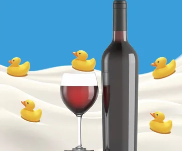 A bottle and glass of red wine with rubber ducks on a white surface against a blue background.