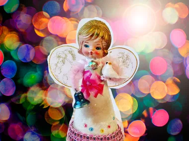 Vintage angel ornament with pink dress, holding a white bird and bell, set against festive, colorful bokeh lights background.