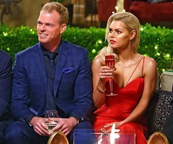 A couple dressed elegantly sit together, holding drinks at a formal event with greenery in the background.