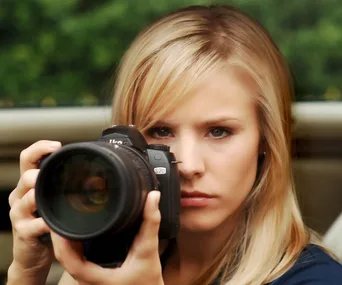 A woman with long blonde hair holds a camera, focused and ready to take a photo.
