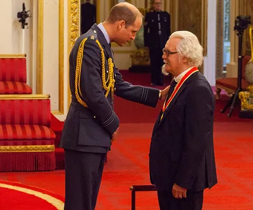 Prince William knighting a man in a formal ceremony at a richly decorated room.
