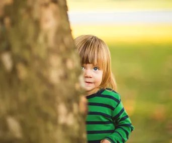 A child with long hair peeks from behind a tree, wearing a green striped shirt, in a park setting.
