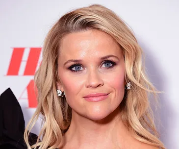 Blonde woman smiling with minimal makeup, wearing earrings, posed against a backdrop with red text.
