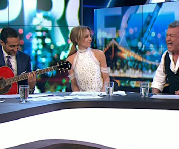 Panel of three people, a man playing guitar, a woman smiling, and another man singing passionately on a TV show set.