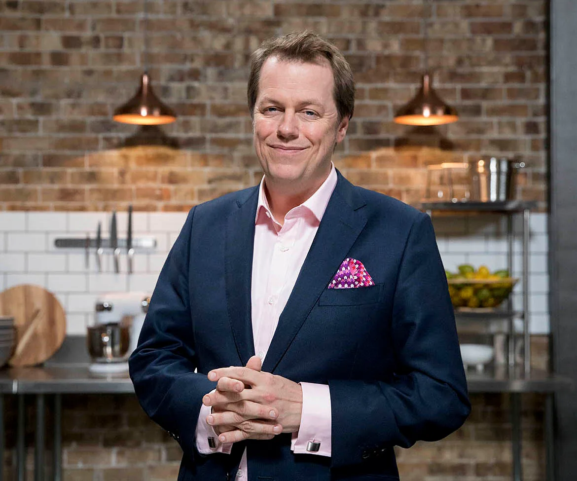 Man in a suit with pink shirt and patterned pocket square, standing in an industrial-style kitchen setting.
